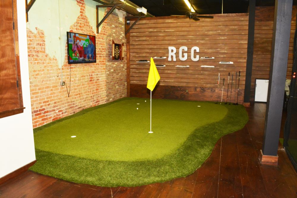 Custom indoor putting green with artificial turf, featuring multiple practice holes and a flag inside a modern recreational space with brick and wood walls.