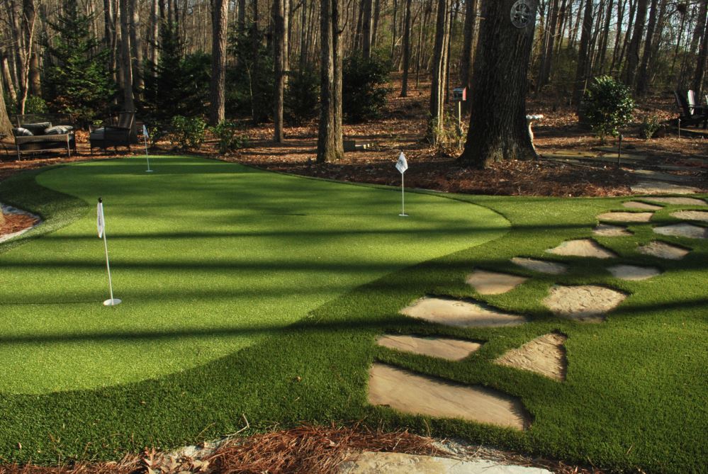 Artificial putting green with multiple practice holes and flags, bordered by a stone walkway, set in a wooded backyard