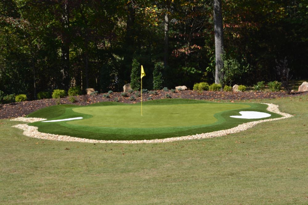 Backyard synthetic turf putting green with flagstick and practice hole, featuring fringe grass, decorative stone edging, and landscaped surroundings.