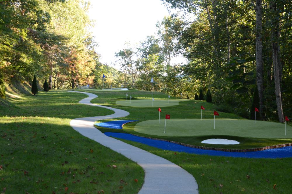 Outdoor synthetic turf putting green course with multiple practice greens and flagsticks, connected by winding walkways and set within a wooded landscape.