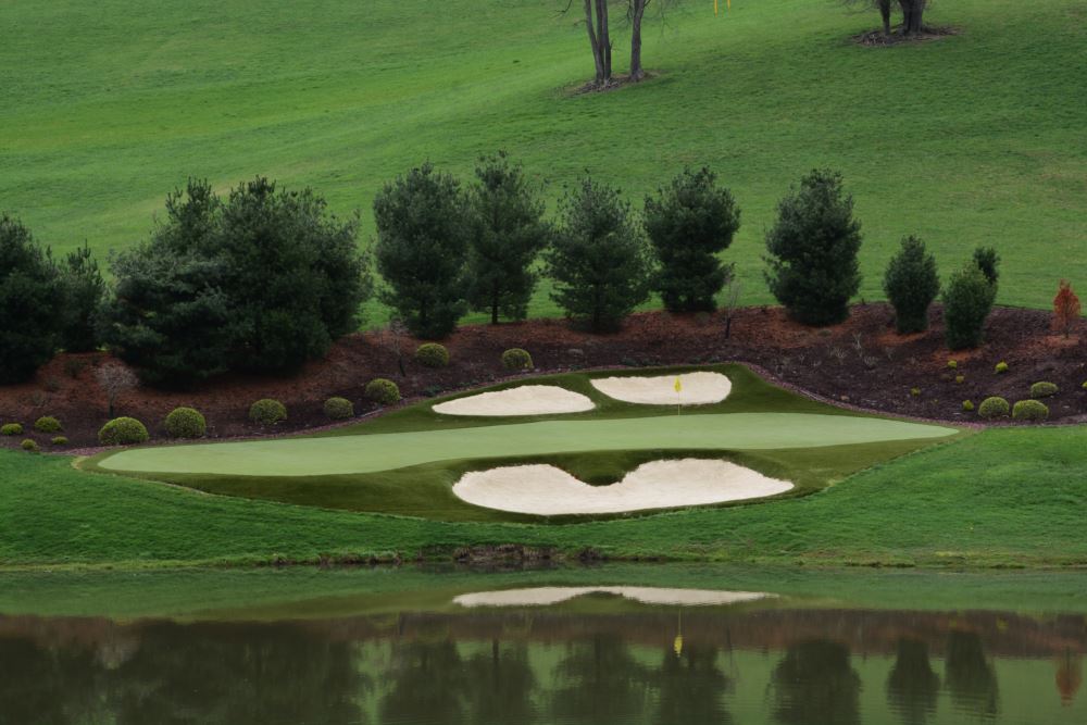Professionally designed golf greens featuring a smooth putting surface with sand bunkers and flagstick, surrounded by manicured landscaping and reflecting over a water feature.